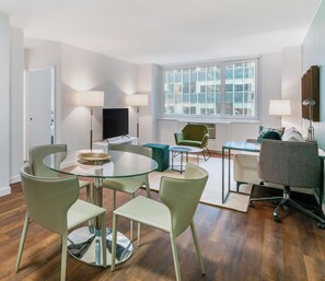 Un salon moderne avec une table à manger au plateau en verre, des chaises vertes, un téléviseur à écran plat et une grande fenêtre donnant sur la ville.