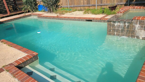 A rectangular swimming pool with clear water, surrounded by a brick border and a stone waterfall feature.