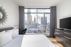 A modern hotel room with a large window offering a city skyline view, a bed with white linens, a black TV on a dark dresser, and a stylish wall mirror.