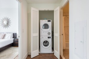 A laundry room with stacked front-loading washers and dryers, a bed with a nightstand, and a wall-mounted mirror.