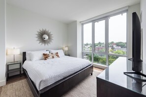 Un dormitorio con un ventanal grande, una cama con ropa de cama blanca, una mesita de noche con lámpara y un espejo fijado a la pared.