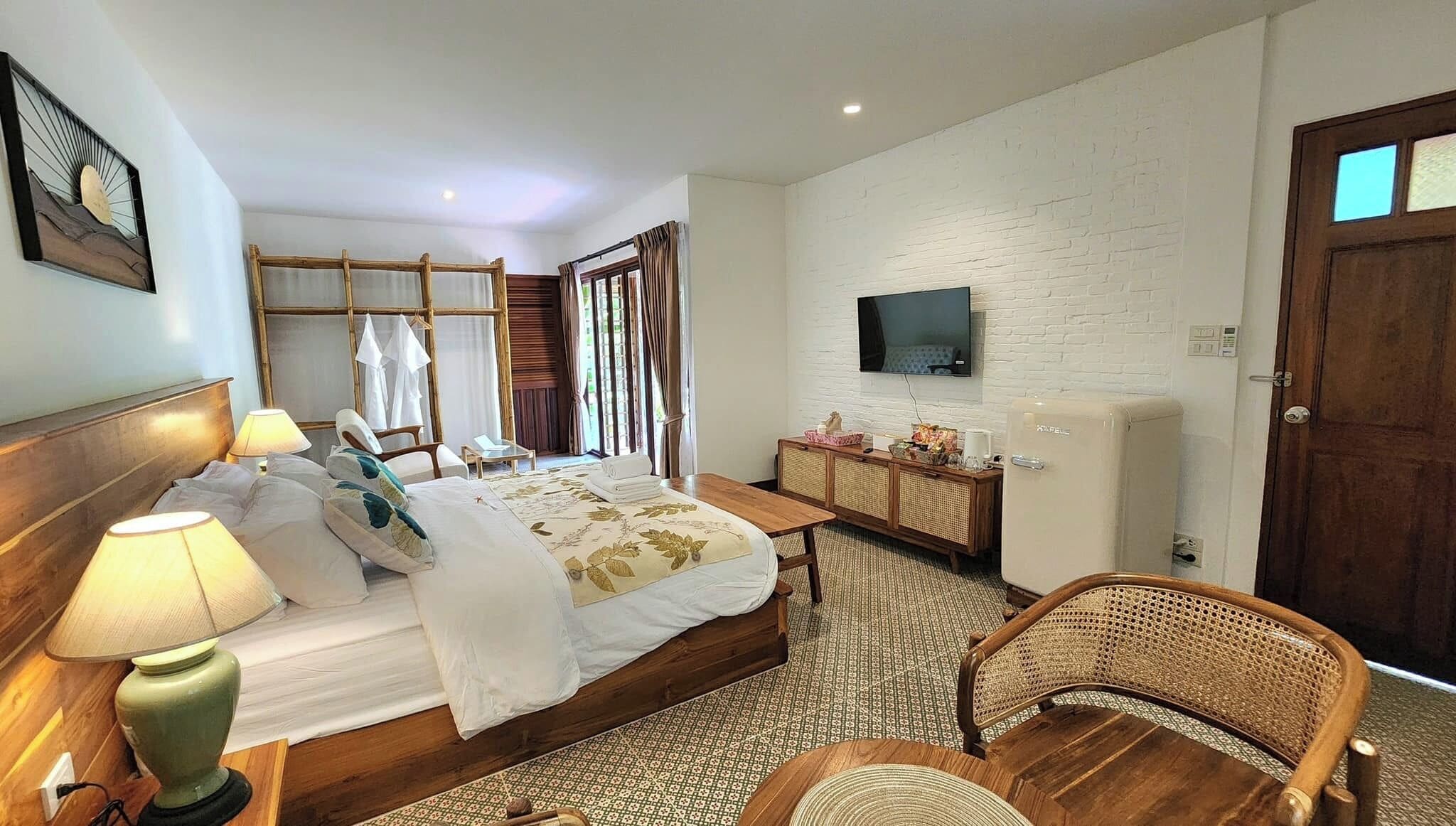 A hotel room with a bed, bedside lamps, a television, a minibar, and a wicker chair.