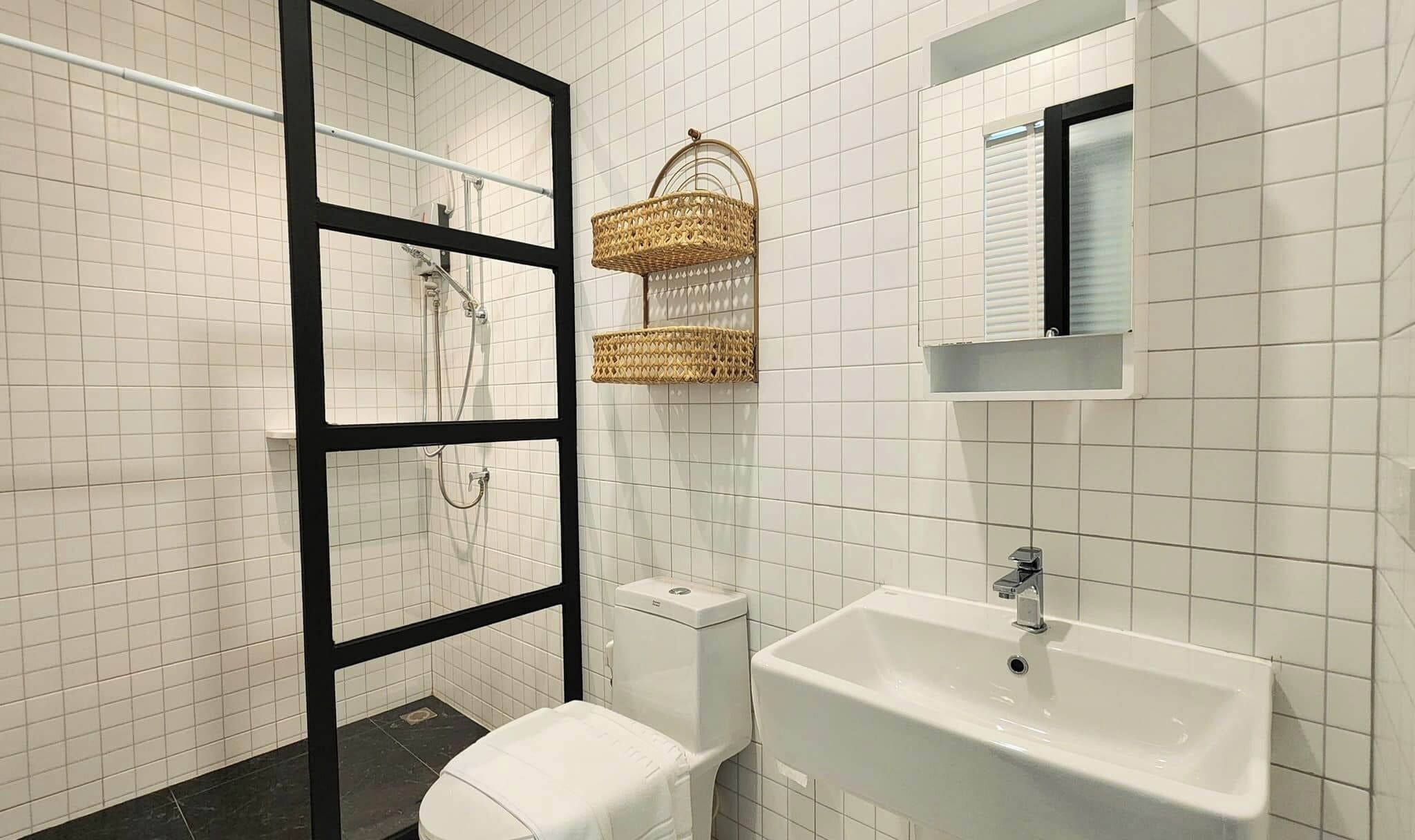 Un baño con paredes de azulejos blancos, cabina de ducha con marco negro, inodoro blanco, lavamanos blanco con grifo cromado y dos cestas tejidas en la pared.