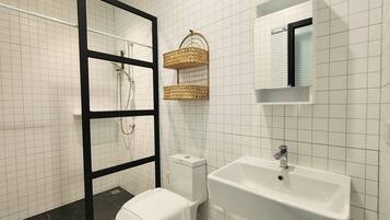 Un baño con paredes de azulejos blancos, cabina de ducha con marco negro, inodoro blanco, lavamanos blanco con grifo cromado y dos cestas tejidas en la pared.