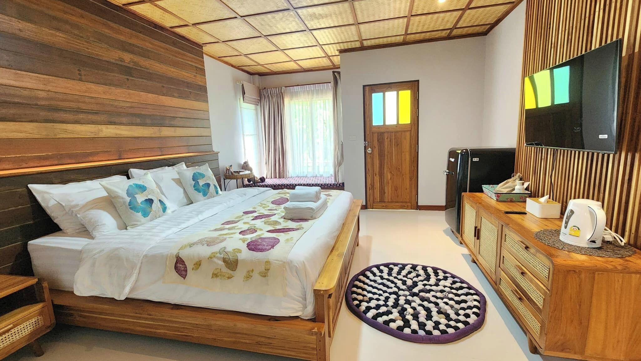 Un dormitorio con cabecera de madera, cama con ropa de cama floral, alfombra redonda, cómoda de madera con televisor y hervidor, y una puerta que da a otra habitación.