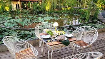Une terrasse en bois, agrémentée de chaises et d’une table en métal peintes en blanc, disposées pour un repas, et entourée d’un étang où flottent des nénuphars et où s’élèvent des palmiers.