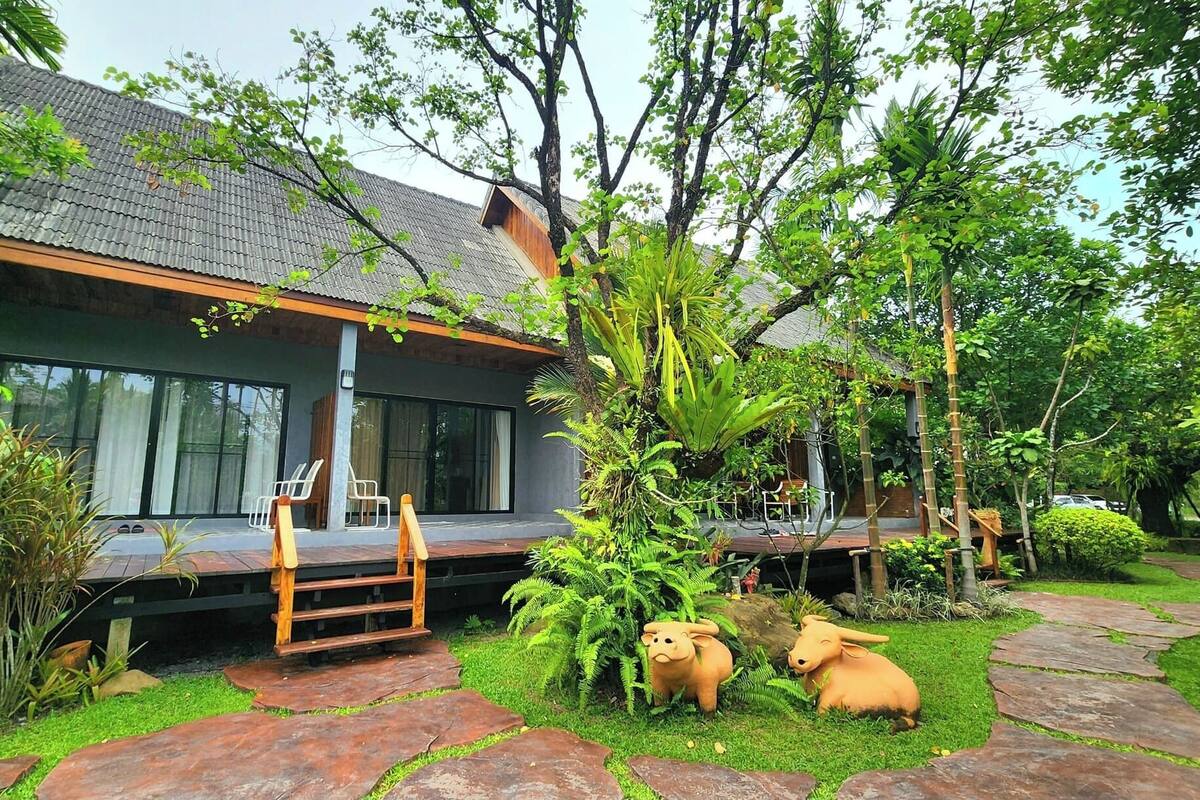 Una casa moderna con un ventanal grande, una terraza de madera y un jardín con plantas tropicales y una estatua de un cerdo.