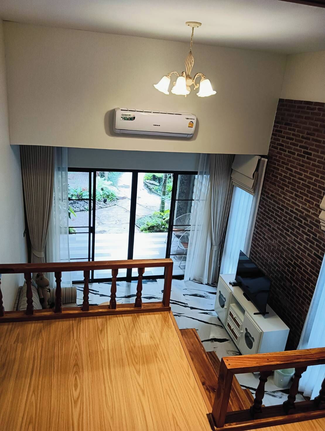 Une pièce spacieuse dotée d’un grand plancher en bois, d’un ventilateur de plafond, d’un lustre moderne et offrant une vue sur un jardin grâce à de grandes fenêtres.