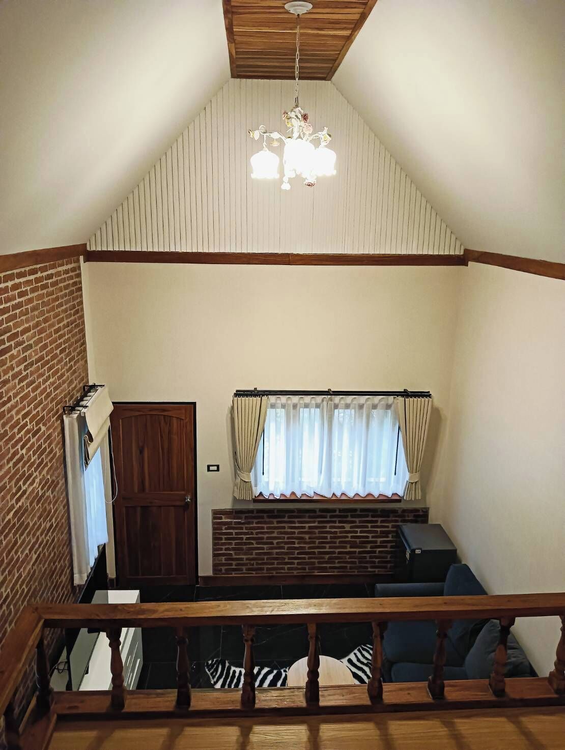 A loft-style room with a sloped ceiling, a wooden staircase, a brick wall, a window with curtains, and a hanging light fixture.