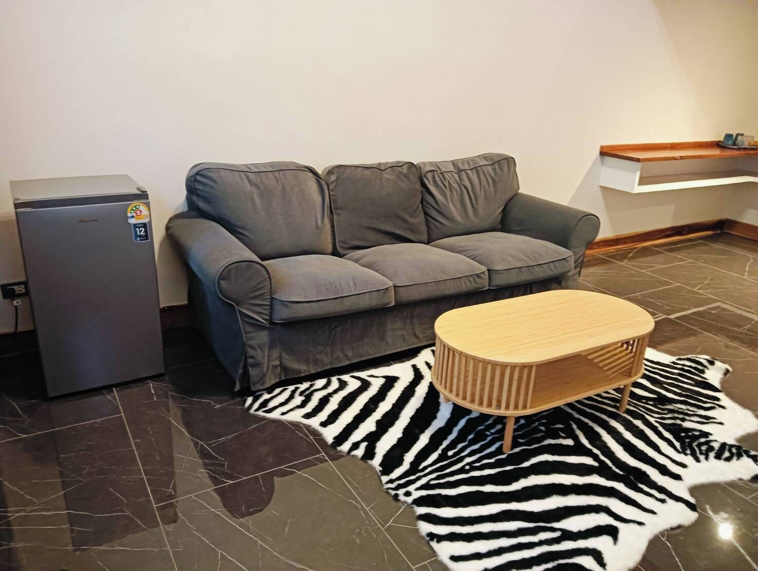Een woonkamer met een grijze bank, een klein houten bijzettafeltje, een tapijtend met zebraprint en een koelkast.