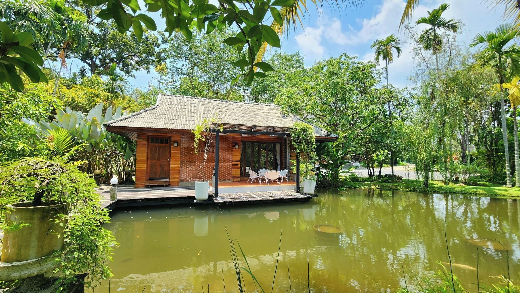 Una casa flotante con revestimiento de madera, techo de tejas y una zona de estar al aire libre cubierta con sillas. Está rodeada de abundante vegetación y una masa de agua tranquila.