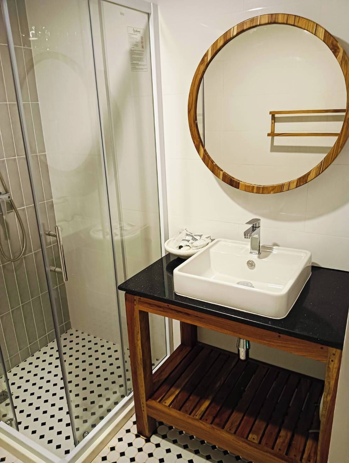 Een badkamer met een glazen douchecabine, een witte wastafel met houten opbergmeubel en een ronde spiegel met houten lijst.