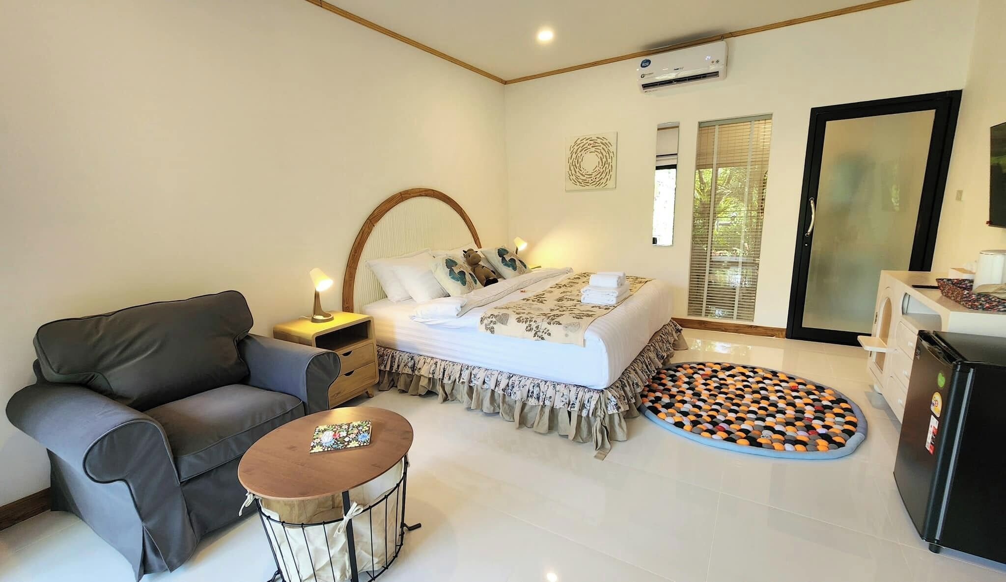 A bedroom with a bed, a sofa, a small table, an air conditioning unit, and a refrigerator.