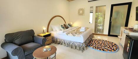 A bedroom with a bed, a sofa, a small table, an air conditioning unit, and a refrigerator.