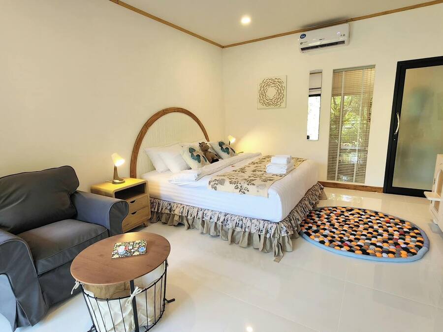A bedroom with a bed, a sofa, a small table, an air conditioning unit, and a refrigerator.