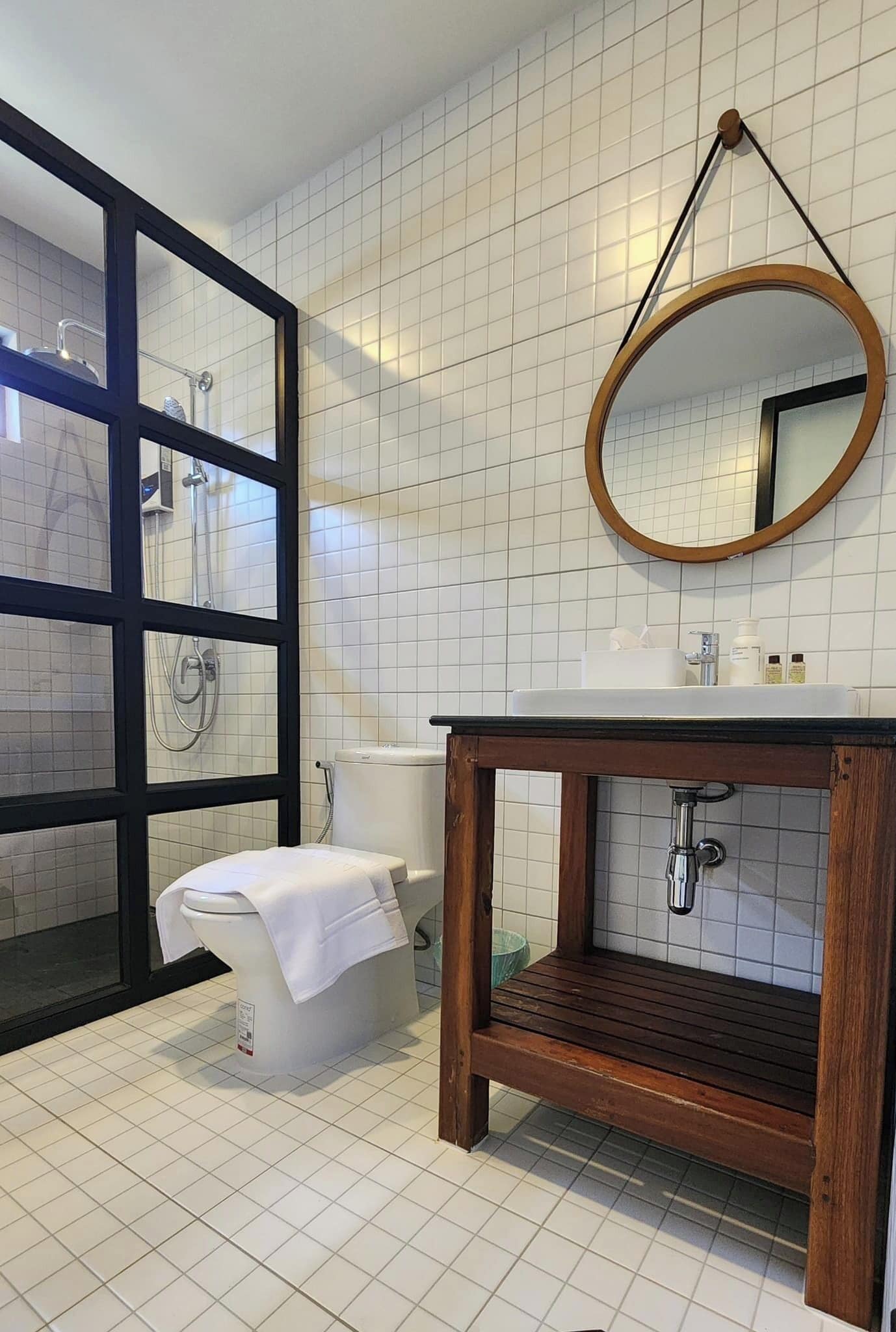 A bathroom with a white tiled floor, a wooden vanity with a sink, a toilet, and a shower area visible through a glass door.