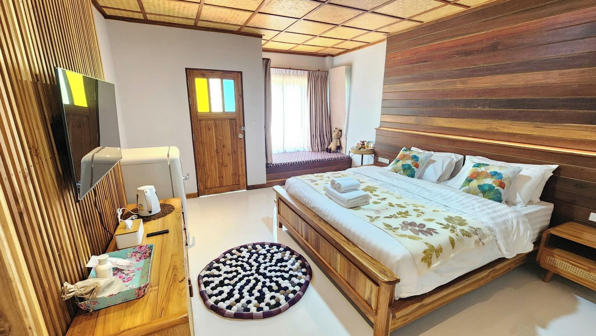 A bedroom with a wooden bed, a bedside table, a round rug, a mirror, a television, and a door leading to another room.