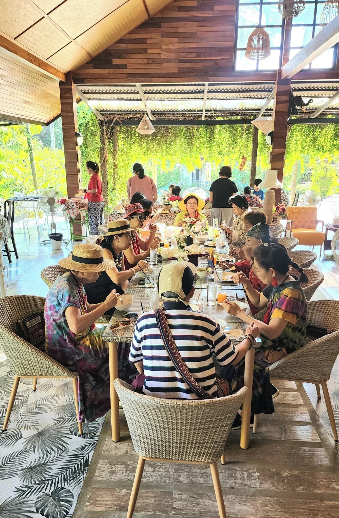 A group of people dining in a covered outdoor area with wicker furniture and a wooden ceiling.