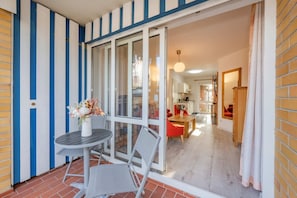 Un balcón con una mesa y sillas, y un ventanal corredero que da a un espacio interior con comedor y cocina.