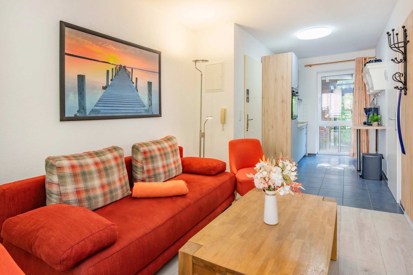 A modern living room with a red sofa, a wooden coffee table, and a painting of a sunset over a pier.