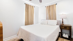 A bedroom with a bed, a ceiling fan, a nightstand with a lamp, and curtains.