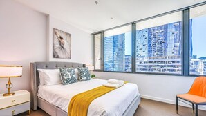 Habitación de hotel moderna con una cama grande, una mesita de noche, una silla y vistas a la ciudad a través de amplios ventanales.
