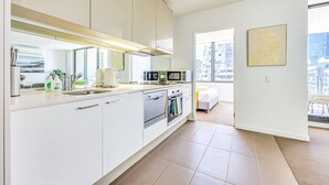 Cocina moderna con armarios blancos, horno integrado y microondas. Hay un comedor con mesa y sillas, y un ventanal con vistas a la ciudad.