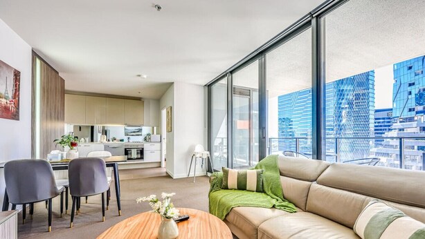 Una sala de estar moderna con sofá, mesa de centro y sillones, una cocina al fondo y ventanas amplias con vistas a la ciudad.