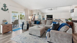 A spacious living room with a large sectional sofa, a coffee table, and a TV. There is a dining area with a table and chairs, and a kitchen area with cabinets and a countertop.