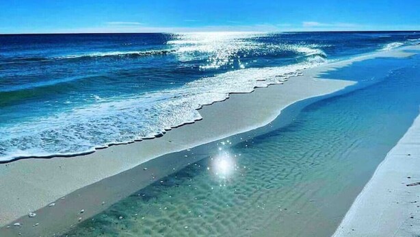 A beach with clear water, a sandy shore, and a bright sun in the sky.