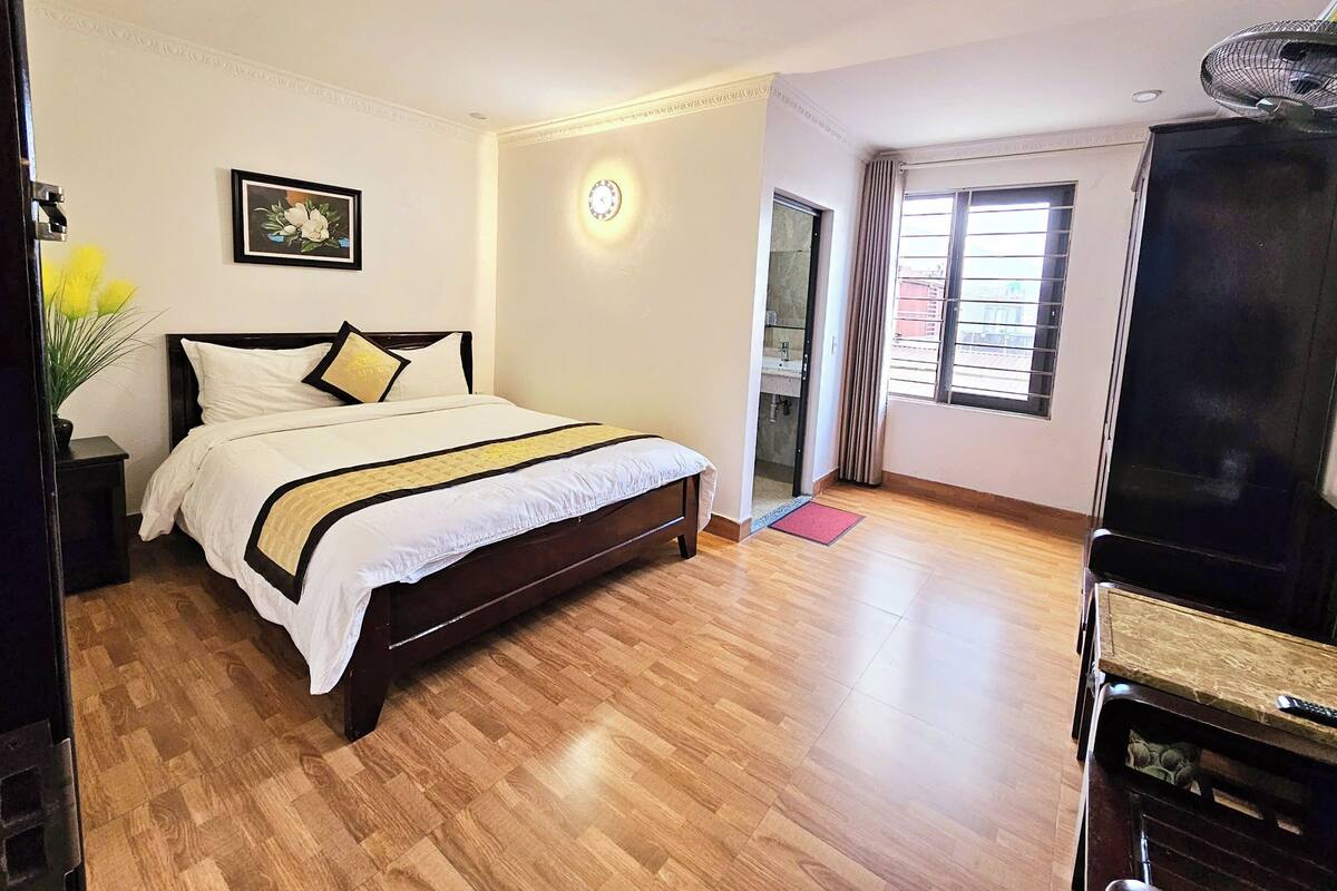 A hotel room with a large bed, a bedside table, a TV stand, a window with curtains, and a wall clock.