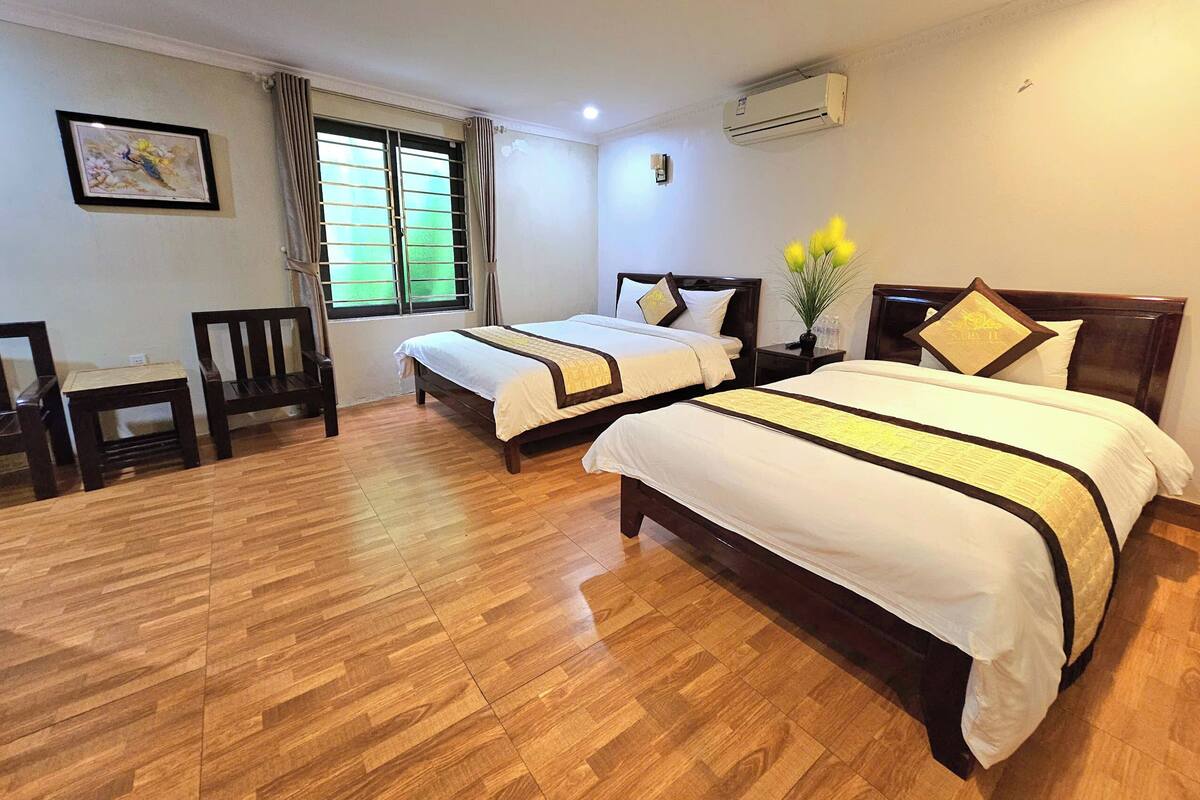 A hotel room with two beds, wooden flooring, a desk, and a window with curtains.