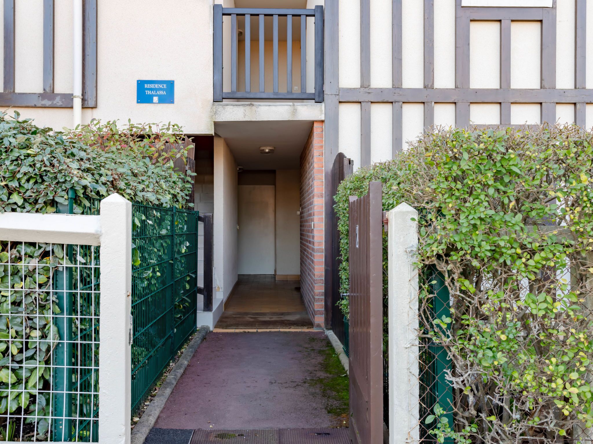 Un étroit sentier mène à l’entrée d’un bâtiment, bordé de clôtures et de feuillages verts. Un panneau bleu indique la présence d’une résidence.