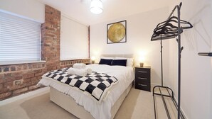 Un dormitorio con pared de ladrillo, cama con manta a cuadros, lámparas de noche y perchero.