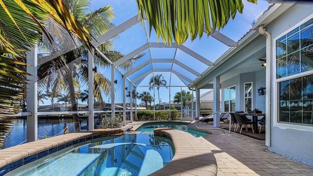Zona de piscina con cerramiento de vidrio, palmeras y un patio cubierto con mobiliario exterior.