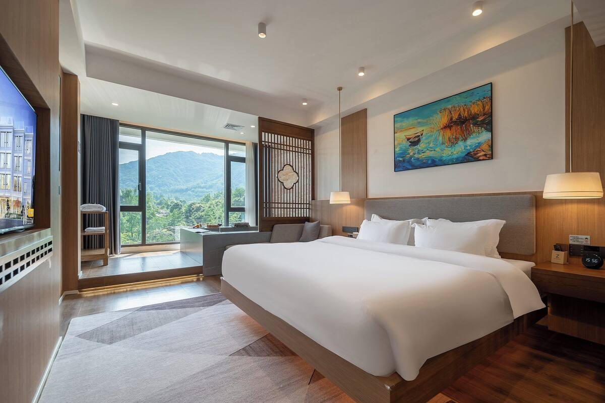 Habitación de hotel moderna con una cama grande, un escritorio, una silla y vistas a montañas desde la ventana.