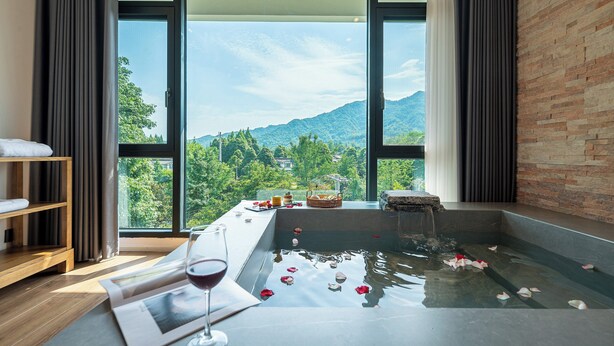 A room with a large window, a bathtub with rose petals, a wooden shelf, and a glass of wine. - Z HOTEL - Qingchengshan (Chengdu)