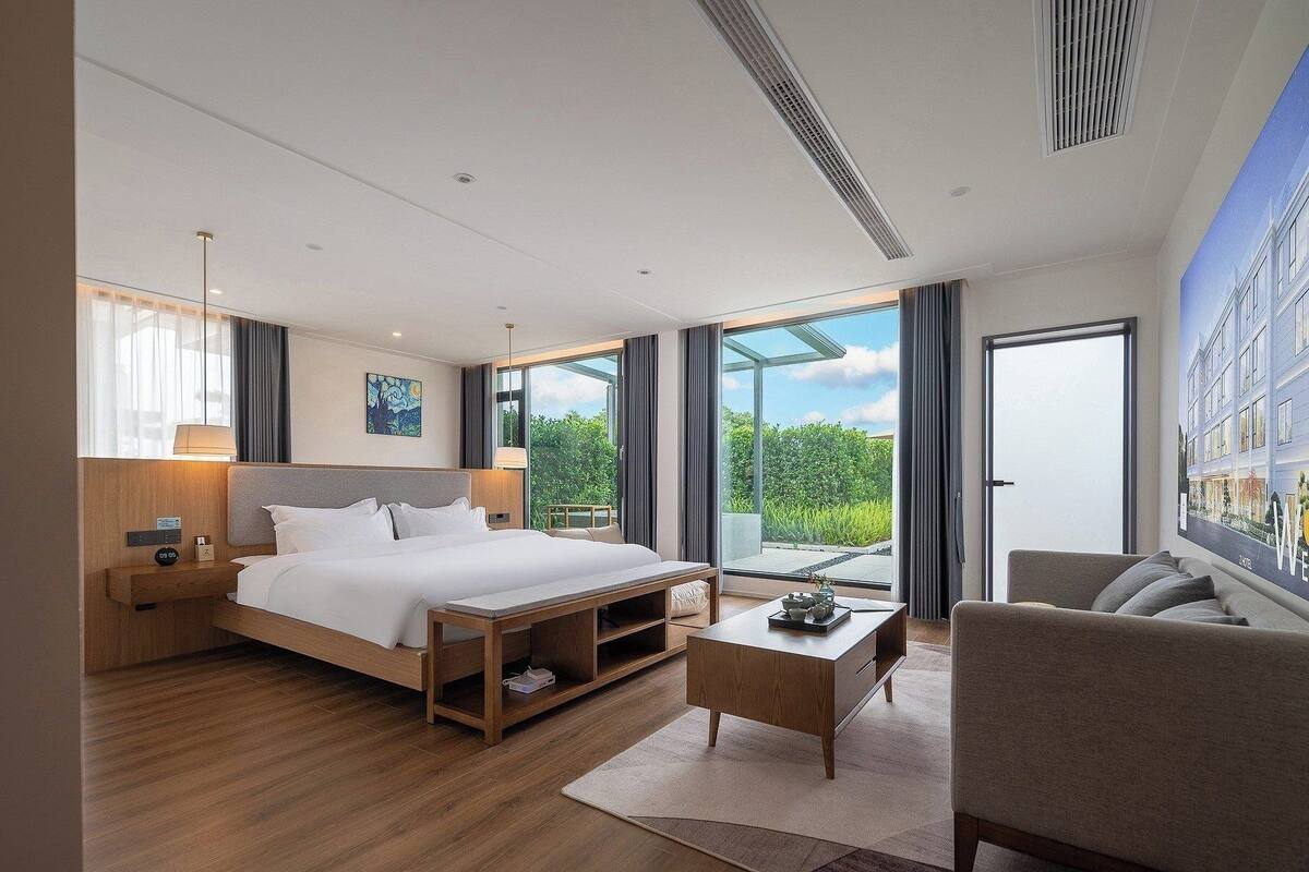 Una habitación de hotel moderna con una cama grande, una mesita de noche de madera, una mesa de centro y vistas a la vegetación a través de las ventanas.