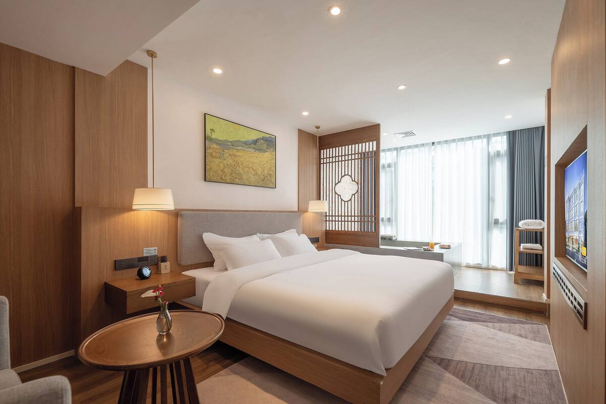 Una habitación de hotel moderna con una cama grande, mesitas de noche, una pequeña mesa redonda con un florero, un cuadro en la pared y un televisor en la pared.