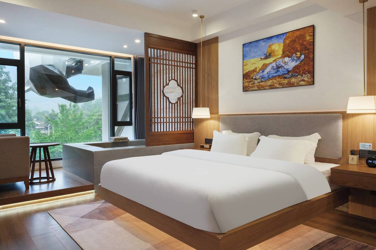 Una habitación de hotel moderna con una cama grande, una zona de estar con una silla y una mesa, vistas a través de un ventanal amplio y un cuadro en la pared.