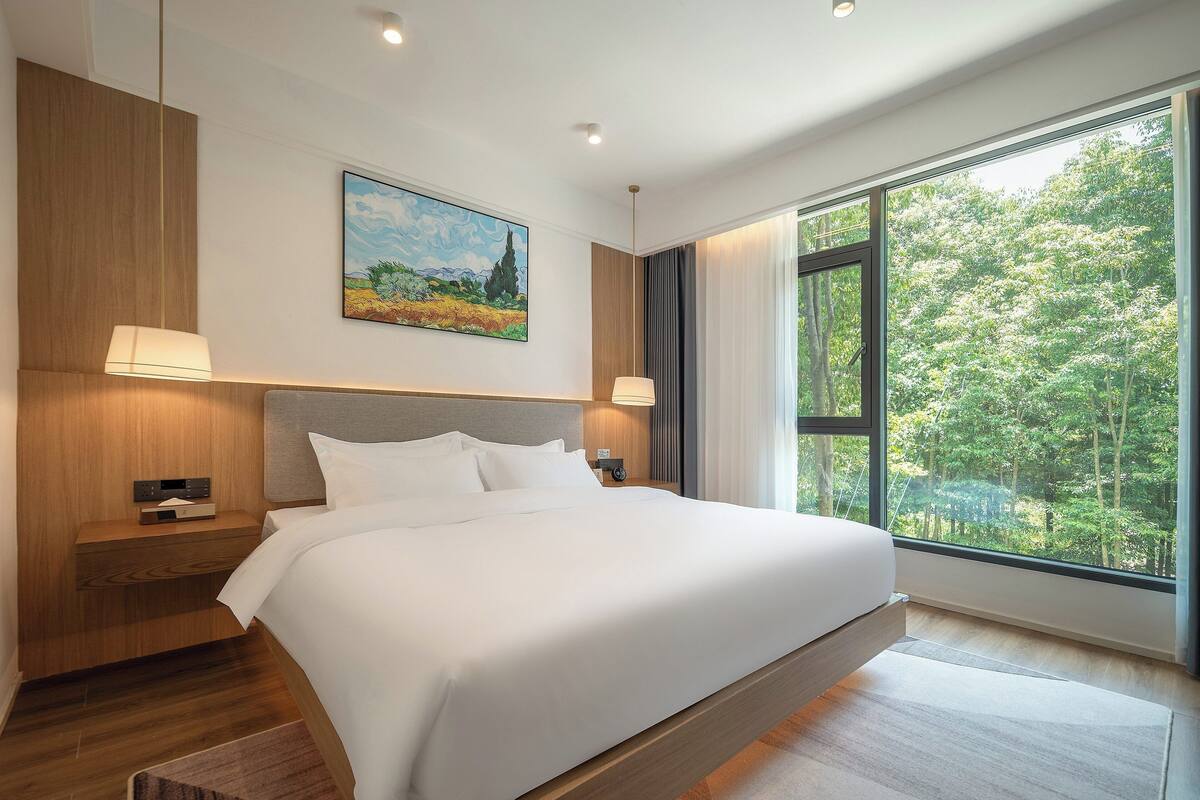 Una habitación de hotel moderna con una cama grande, cabecera de madera, lámparas de noche, un cuadro y un ventanal con vista a un bosque.