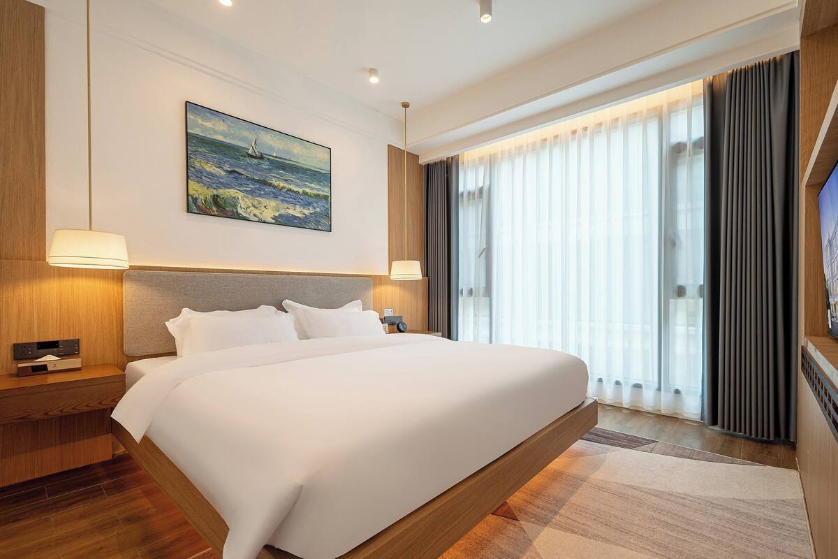 Habitación de hotel moderna con una cama grande, un cuadro de un paisaje marino, un televisor y un ventanal con cortinas.