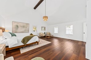 A spacious bedroom with a large bed, a wooden ceiling beam, a hanging light fixture, and a view of the outside through a window.