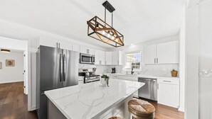 A modern kitchen with white cabinetry, stainless steel appliances, a marble island, and a unique geometric pendant light. - The Grand Haven on the Ashley (North Charleston)