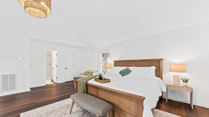A bedroom with a large bed, bedside tables, a bench, a hanging light fixture, and a door leading to another room. - The Grand Haven on the Ashley (North Charleston)