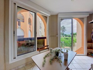 A room with a large sliding glass door leading to a balcony, a dining table with a potted plant, and a view of a building with arches. - Dominica by Interhome (Le Barcarès)