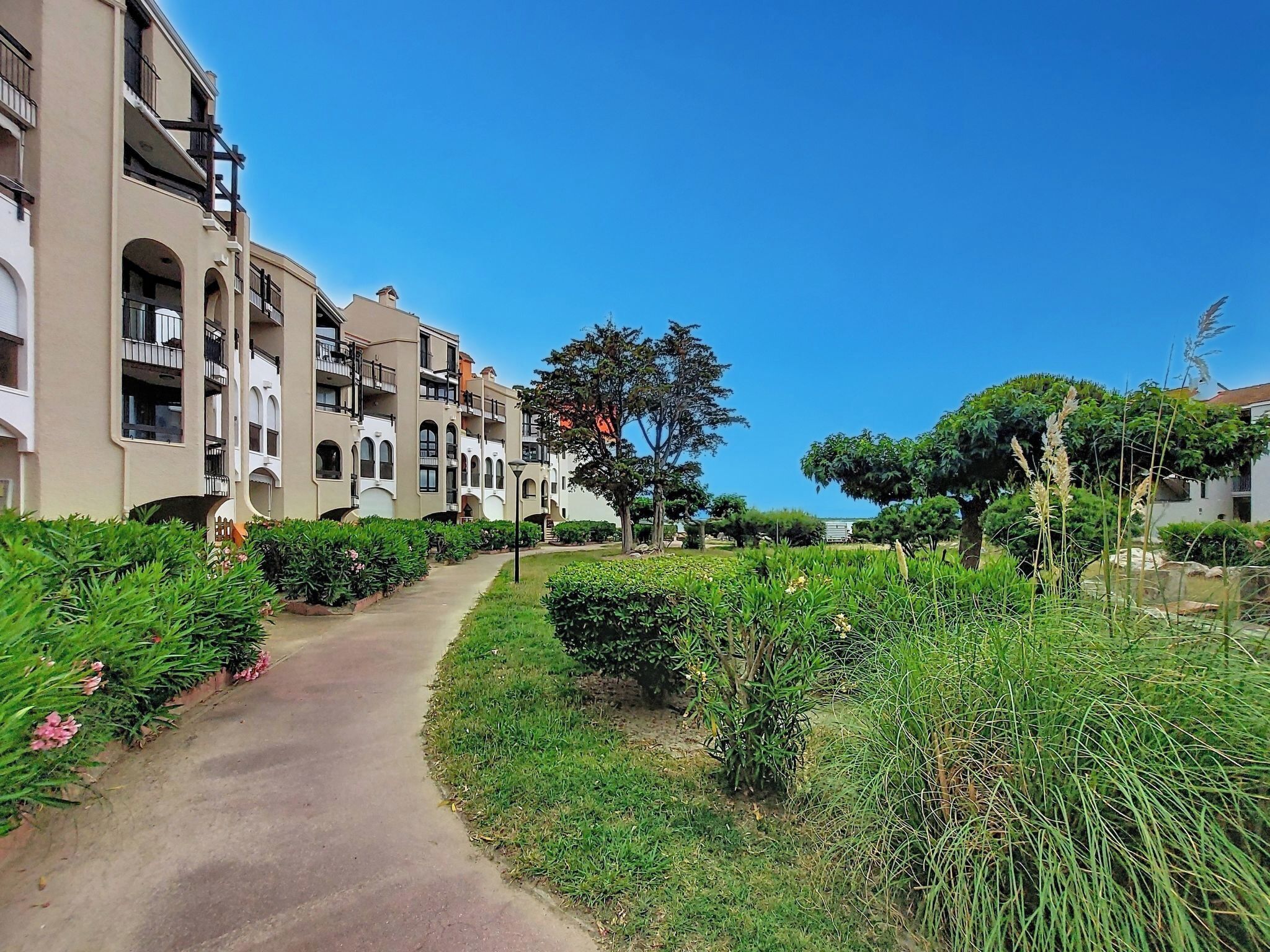 A pathway leading to a multi-story building with balconies, surrounded by well-maintained greenery and trees.