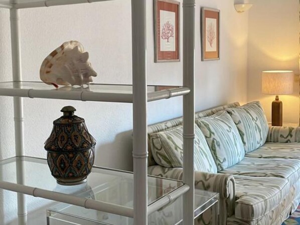A living room with a glass shelving unit, a patterned sofa, and framed artwork on the wall.