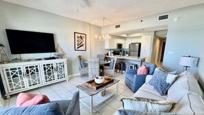 A modern living room with a flat-screen TV, a dining area with a kitchen in the background, and a ceiling fan. - Direct Gulf Front 1/1 Sleeps 6 (Orange Beach)