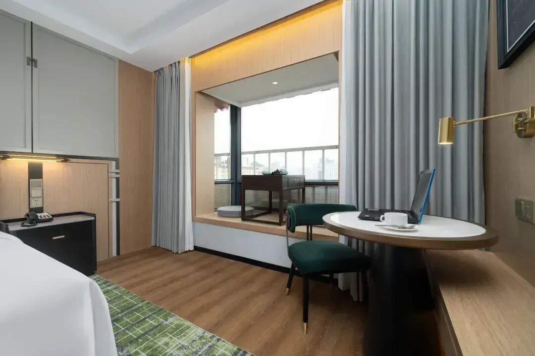 A hotel room with a bed, a desk with a laptop, a chair, a small table, and a window with curtains.